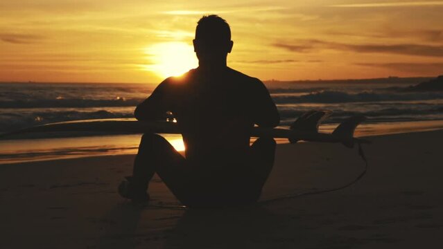 Surfer with surfboard sitting on beach at sunset