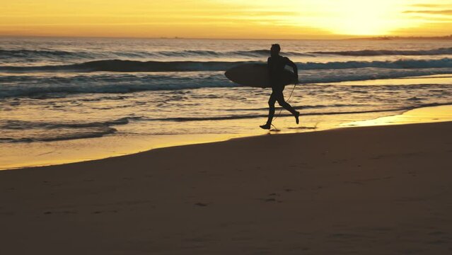 Surfer with surfboard running by ocean at sunset