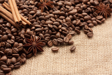 Roasted coffee beans are scattered on sackcloth with star anise and cinnamon sticks. There is copyspace for your text.