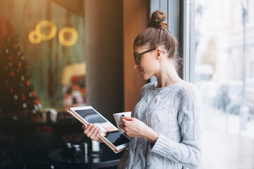 Young girl looking at tablet and smiling in cafe with big window. She has beverage in hand