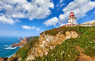 Fototapeta na wymiar Lighthouse at Cape Roca (Cabo da Roca), most western point of Europe at coast of Atlantic Ocean in Portugal. Rocks at coastline and blue sky with clouds over skyline. Picturesque summer landscape.