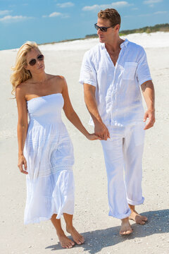 Sad thoughtful man and woman romantic couple in white clothes and sunglasses holding hands walking on a deserted tropical beach with clear blue sky, relationship issues concept.