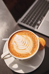 A cup of coffee in hand next to laptop. A cup of hot cappuccino coffee with cookies and a laptop computer on the table in the coworking space of the coffee shop. Online business, work in cafe