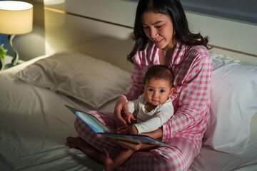 mother read story in the book for infant baby on bed at night
