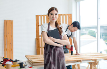 Asian professional female carpenter in apron smiling holding notes on clipboard while male...
