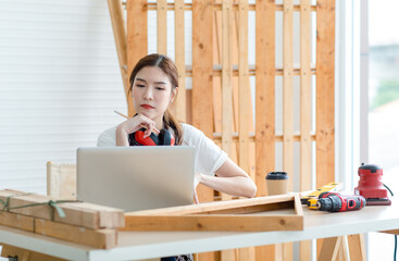 Asian professional thoughtful female carpenter worker staff with earphones in apron sitting holding pencil thinking ideas via laptop notebook computer on workbench in interior home building workshop