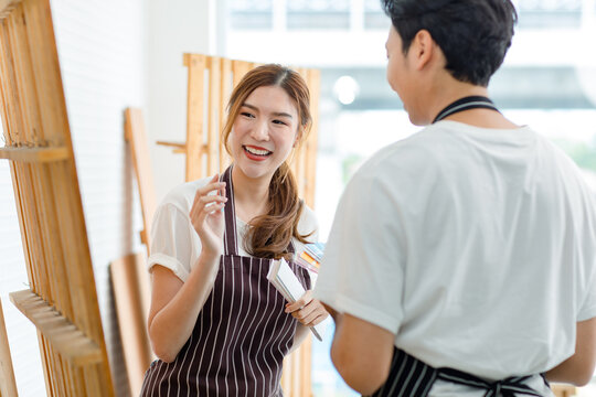 Asian professional female carpenter worker lover in apron smiling showing pointing chosen light blue color chart pattern to male boyfriend couple colleague helping choosing interior home colour