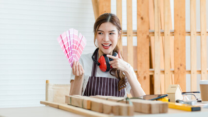 Asian professional cheerful female carpenter worker staff in apron with earphones sitting smiling showing colorful color chart pattern via video call on laptop computer in home building workshop