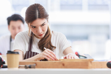 Obraz premium Asian professional focused female carpenter worker staff in apron sitting holding using measuring tape measure wooden sticks on workbench while male lover colleague working on blurred background