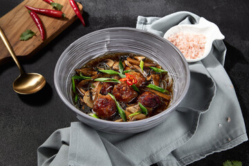 Horizontal frame of a delicious Asian soup enriched with mushrooms and meatballs, surrounded by vibrant ingredients on a black backdrop. The scene is accentuated by a stylish textile napkin.