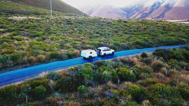 Drone follow shot. Vehicle towing camping trailer down dirt road with sun rise in the snowy mountains of New Zealand southern alps.