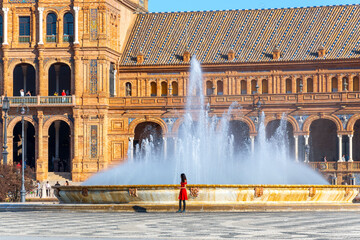 An unidentifiable woman in a red dress stands in front of the main water fountain at the Plaza de...