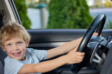 Cute little boy behind the wheel of an expensive car holds the steering wheel, happily looks into the camera. Fulfilling a child's dream of sitting behind the wheel in a real car