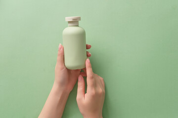 Two hands of female are holding a green cosmetic bottle unbranded isolated on a green background. Minimal concept for advertising cosmetic with hand model. Front view, space for design