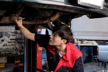 Professional young Asian male motor mechanic inspects undercarriage of electric car(EV) lifted by forklift jack for repair at garage, automotive maintenance service works industry occupation business.