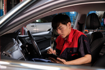 One young expert Asian male automotive mechanic technician checking a maintenance list with tablet in car interior at garage. Vehicle service fix and repair works, industrial occupation business jobs.