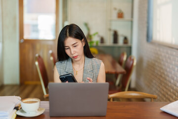Close-up young Asian woman sitting on mobile phone, while resting eyes from work, looked at laptop screen on desk in front expression looked cheerful happy, as typed read text on the screen.