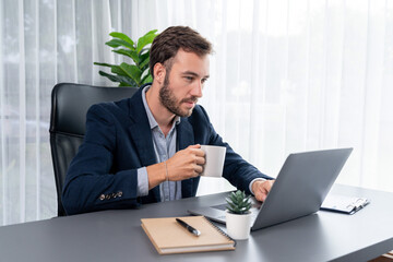 Businessman in black suit working on laptop at his workspace desk. Smart executive researching financial data and planning marketing strategy on corporate laptop at modern workplace. Entity