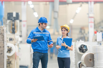 An American male engineer manager and a female employee walk to inspect the order in a large plastic and steel factory. Helmets, uniforms and listnotes, walkie-talkies, machines working around.