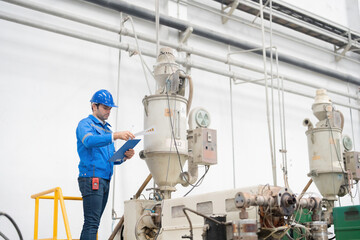 American male engineer Standing on a working machine Holds a listnote, checks the materials list, wears a uniform, helmet, and has a walkie-talkie. in a large plastic and steel industry