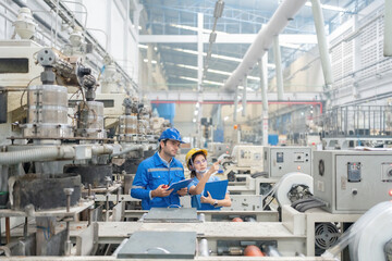 American male engineer manager and female employee working together pointing to destination There is a working machine Wear helmets, uniforms and listnotes in large plastic and steel factories.