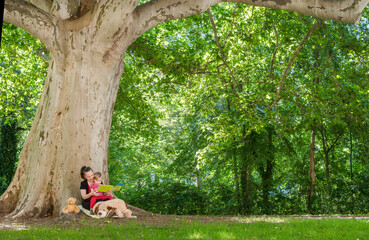 Mother reading a book with her cute little child under an old plane tree in the park. Funny and educative outdoor activity for children, in sunny summer day