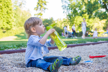 Cute little toddler boy drinking water from a big green bottle in the park, hydrating properly in hot summer day