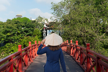 Asian woman is wearing Ao Dai traditional Vietnamese dress and traveling at Red Bridge- The Huc Bridge in Hoan Kiem Lake, this is a lake in the historical center of Hanoi, the capital city of Vietnam
