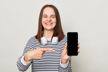 Fototapeta premium Attractive cheerful woman wearing striped shirt standing with headphones over neck isolated over gray background pointing at smartphone with empty display copy space for advertisement