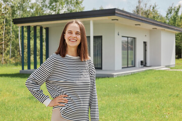 Dark haired smiling standing near building new house on green lawn keeps hand on hip smiling to...