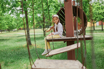 Smiling dark haired little girl wearing casual clothing and baseball cap playing on playground in green park sitting looking at camera enjoying sunny day.