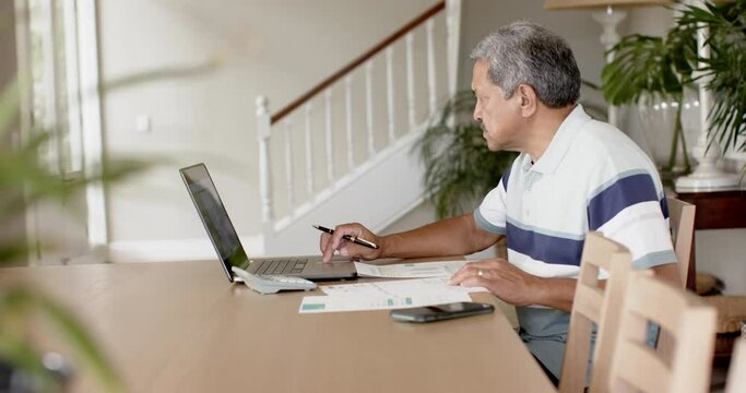 Biracial senior man doing paperwork and using laptop at home, slow motion