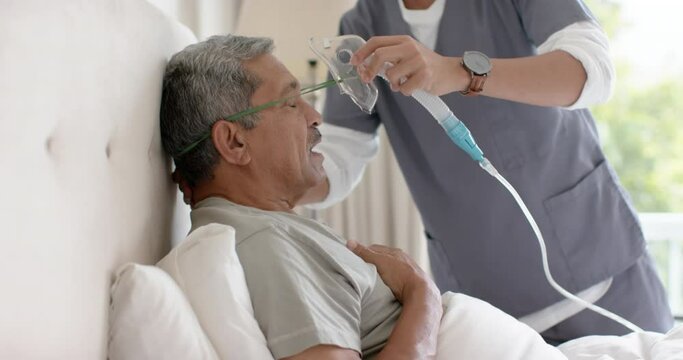 Diverse male doctor helping senior male patient using oxygen mask at home, slow motion