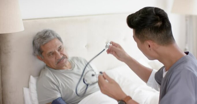 Diverse male doctor testing blood pressure of senior male patient in bed at home, slow motion