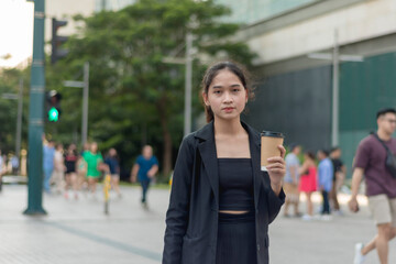 A lovely lady standing outside, in the city wearing a black suit, and black skirt is seriously looking at the camera while holding her coffee. 