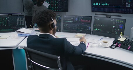 African American trader works at computer with displayed real-time stocks. Coworkers analyze exchange market charts on big screens at background. Trading and investment concept. Back view. Dolly shot.