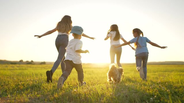 Happy children run together on green field of park at sunset. Rear view of children running across field. crowd of children run along green grass in summer park. Happy childhood.