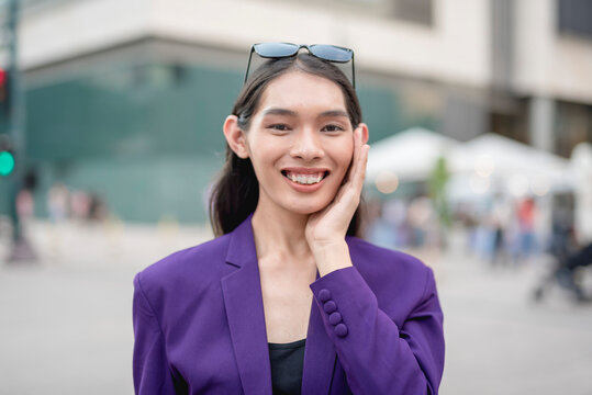 A close- up shot of an asian transwoman standing outside wearing a purple suit and sunglasses on her head, widely smiling while her palm is on her cheek. Building, and tents in the background.