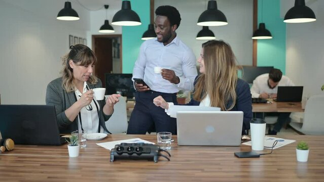 Real time shot of diverse smiling women with man having coffee and discussing papers while coworking in modern office