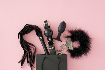 Sex shop of BDSM-inspired erotic toys. Top view photo of feather handcuffs, leather whip, ball gag, anal plug, black shopping bag on pastel pink background - obrazy, fototapety, plakaty