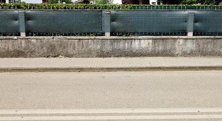 House fence made of concrete low wall, green tarpaulin and hedge. Cement tile sidewalk and street...