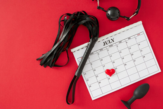 Theme of BDSM Day for intimate exploration. Top view arrangement of calendar marking special date, anal plug, gag, leather whip, heart on red background with blank space for text or promotion