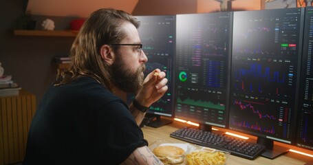 Concentrated trader watches real-time stocks, exchange market charts on multi-monitor computer workstation, types on keyboard. Man works remotely in investment at home office. Cryptocurrency trading.