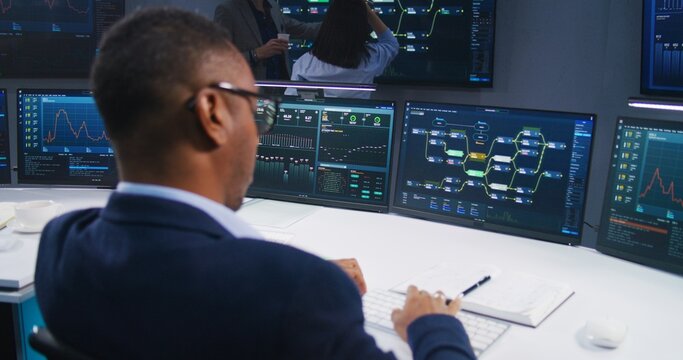 Software programmer sits in front of computer with blockchain network and data server in monitoring office. Team of IT technical specialists work with live analysis charts on big digital screens.