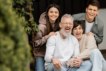 Positive children looking at camera while hugging middle aged parents holding hands and sitting on porch of house during parents day celebrating , parent-child relationship concept
