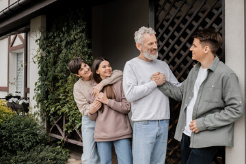 Fototapeta premium Positive middle aged father shaking hands with young son near family during parents day celebration near porch of house at backyard, parent-child relationship concept, special occasion