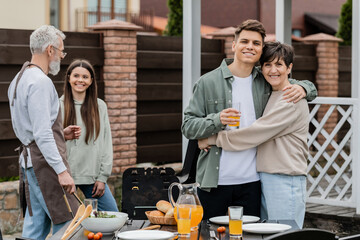 happy parents day, modern parenting, cheerful middle aged mother hugging young adult son holding glass of orange juice, father and daughter preparing food on bbq grill, summer, backyard, celebration