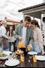 celebration of parents day, modern parenting, happy middle aged mother leaning on young adult son pouring orange juice in glass, father and daughter preparing food on bbq grill, summer, backyard