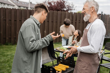 tattooed and bearded father and son preparing food on bbq grill, grilling corn, communication, family bbq party, middle aged, happy parents day, translation of tattoo: harm none do what you will - obrazy, fototapety, plakaty