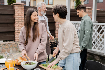 cheerful mother and teenage daughter making salad, father and son preparing food on bbq grill, family house backyard, suburban life, happy parents day celebration, summer, june, candid